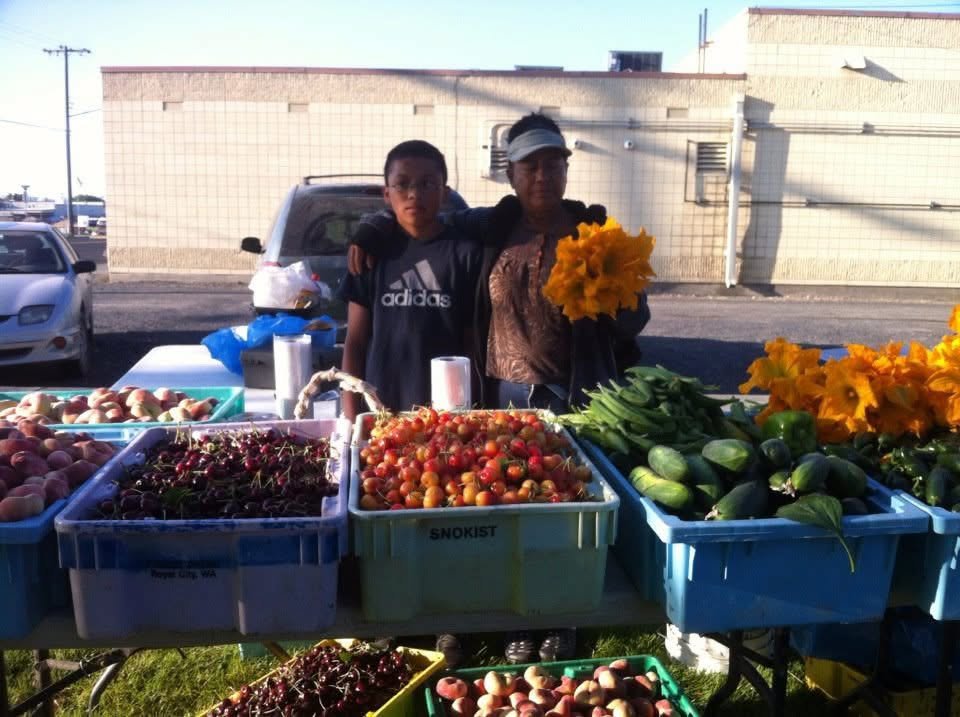 Isaias as a boy next to his grandmother at the family fruit stand. She has one arm around him and holds a bunch of squash blossoms; in front of them are crates of cherries, peaches, and cucumbers.